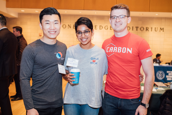 3 people stand facing the camera smiling, all are dressed in jeans and T-shirts bearing logos of tech companies. In the middle is a woman with short dark hair holding a cardboard coffee cup, wearing a grey Tshirt which says Girls who Tech